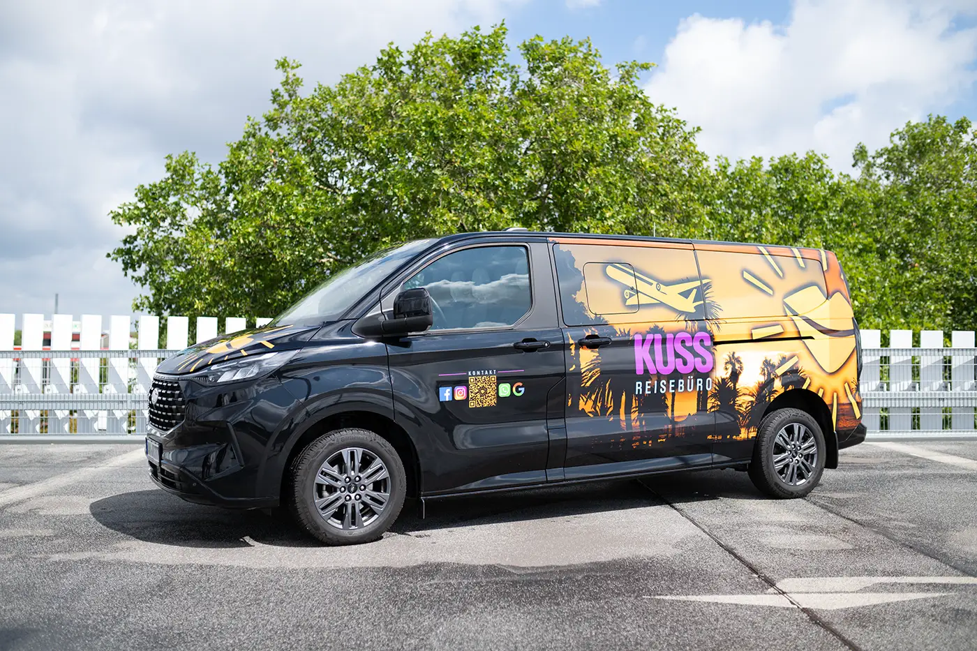 Clickfortravel Schwarzer Van mit bunten Palmenmotiven und Schriftzug "KUSS REISEBÜRO" steht vor weißem Zaun und Bäumen im Freien. Kuss Reisebüro
