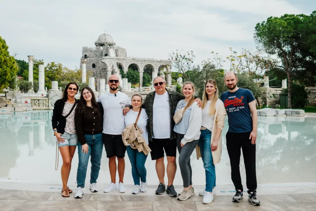Clickfortravel Acht Erwachsene vor einem großen Wasserbecken, antike Steinruinen und viel Grün im Hintergrund, alle lächeln. Kuss Reisebüro